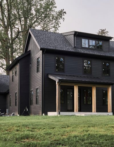 A two-story black house with a covered front porch, multiple windows, and a manicured lawn, surrounded by tall trees.