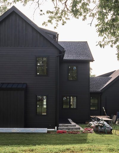 A modern dark-colored house with multiple windows stands amidst trees, surrounded by construction materials and equipment on a grassy yard.